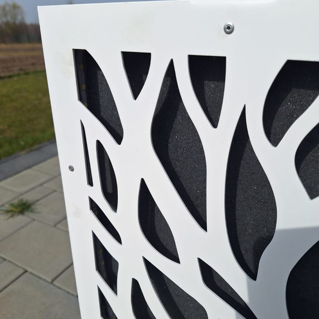 Zdjęcie przedstawia fragment panelu maskującego użytego do zasłonięcia betonowej donicy. Na zdjęciu widoczny jest montaż za pomocą śrubek. Panel jest koloru białego.